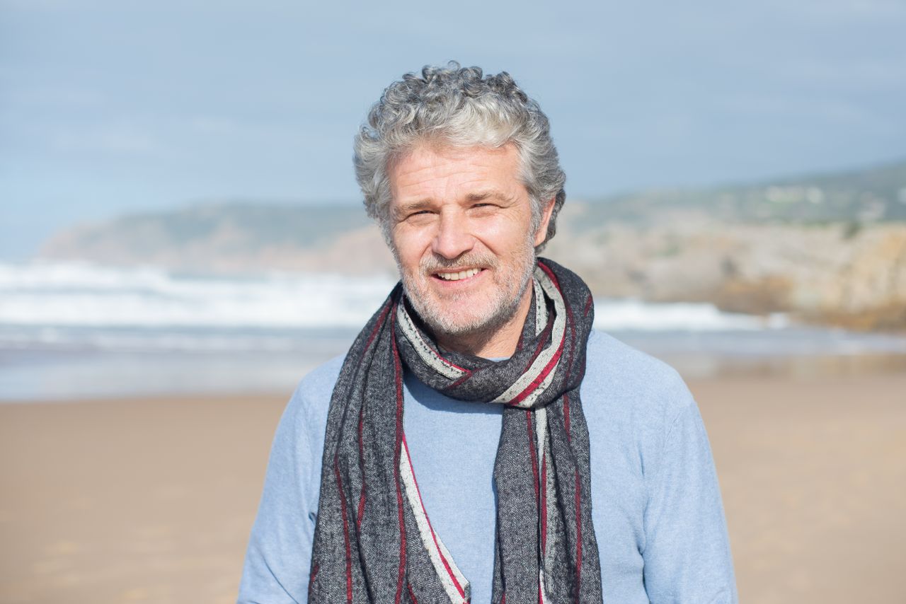 Homme aux cheveux gris souriant, une écharpe autour du cou, au bord de la mer