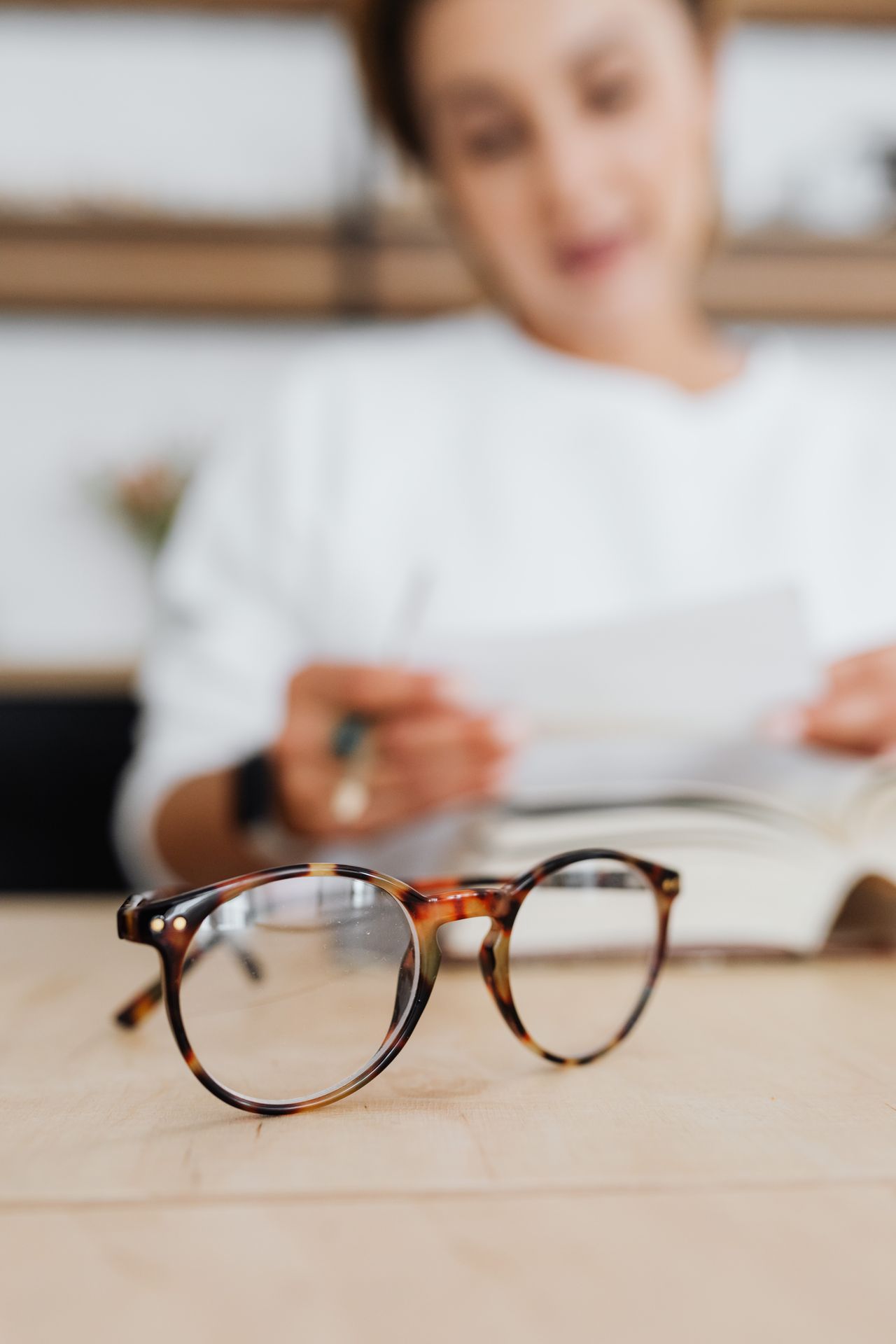 Lunettes posées sur une table, nettes au premier plan, devant une femme floue qui lit